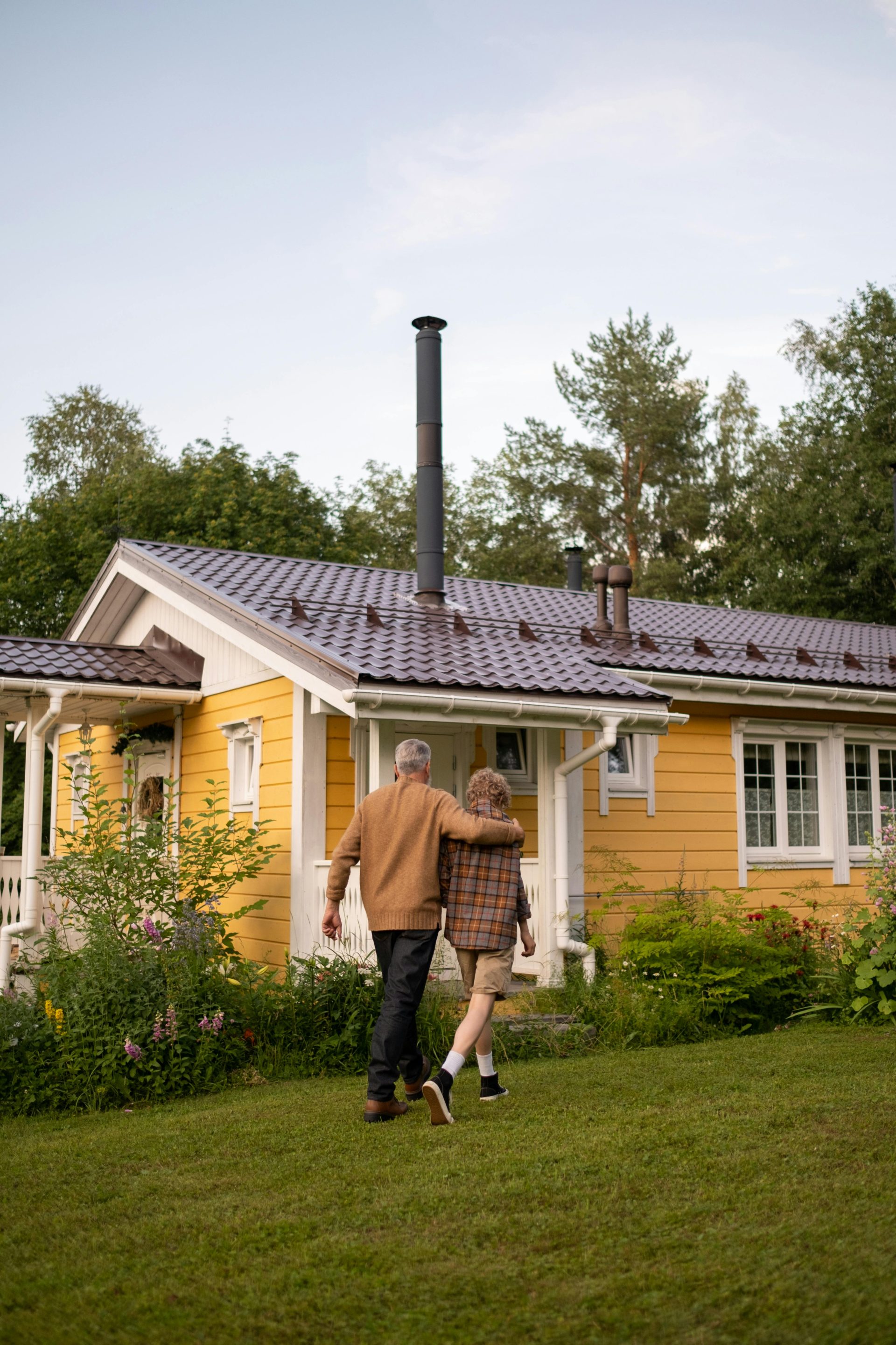 A serene moment of togetherness as a grandparent and child walk by a charming yellow cottage in lush greenery.
